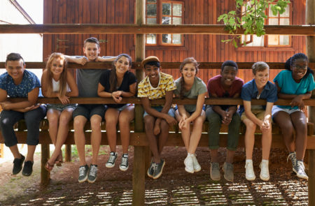 A diverse group of teenagers sit together on a fence, smiling at the camera. Summer Camp Safety.
