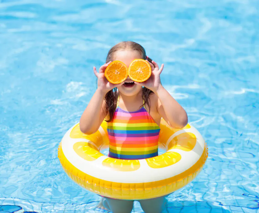 Little girl in swimming pool with inflatable toy ring eating orange. Kids swim on summer vacation. Tropical fruit and healthy snack. Swim aids for child. Kid on colorful float. Beach and water fun.